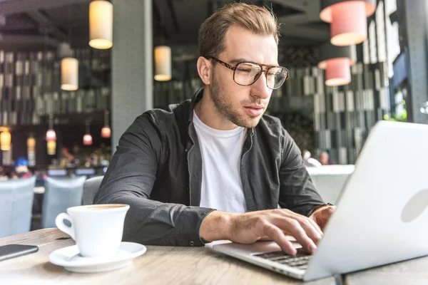 Concentrated young freelancer with cup of coffee working with laptop in cafe — Stock Photo