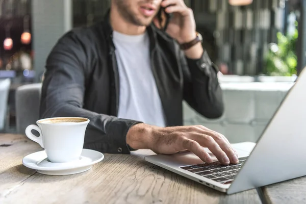 Tiro recortado de jovem freelancer com xícara de café trabalhando com laptop no café e falando por telefone — Fotografia de Stock