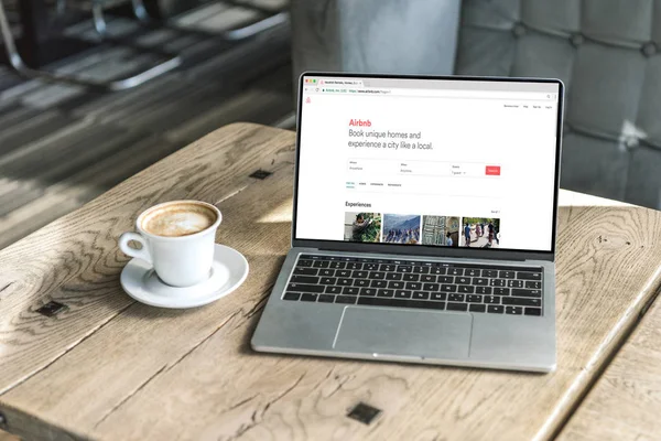 Cup of cappuccino and laptop with airbnb website on screen on rustic wooden table at cafe — Stock Photo