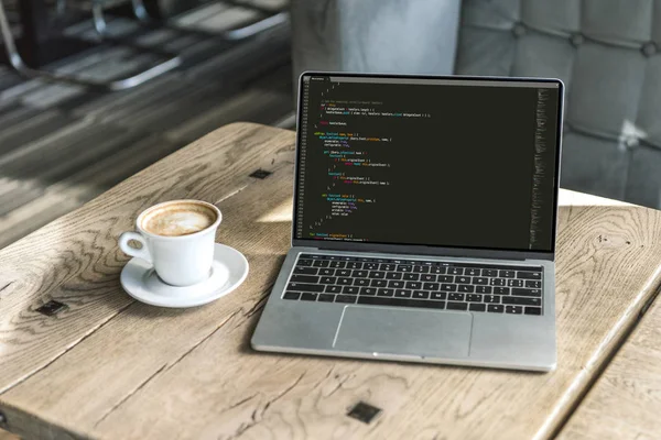 Tasse Cappuccino und Laptop mit Programmcode auf dem Bildschirm auf rustikalem Holztisch im Café — Stockfoto