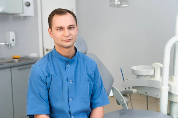 Confiado joven dentista sentado en el consultorio del dentista y mirando a la cámara - foto de stock