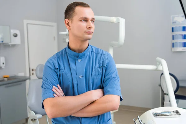 Schöner junger Zahnarzt mit verschränkten Armen, der im Büro wegschaut — Stockfoto