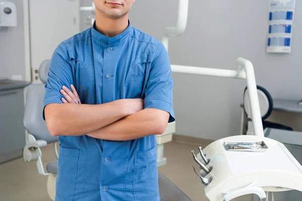 Colpo ritagliato di giovane dentista in uniforme blu con braccia incrociate in ufficio — Foto stock