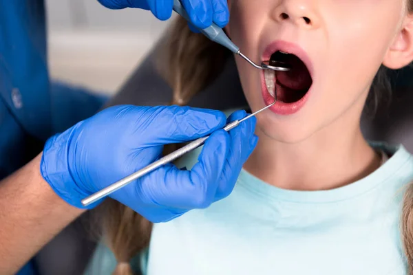 Tiro recortado del dentista con instrumentos que examinan los dientes de un niño pequeño - foto de stock