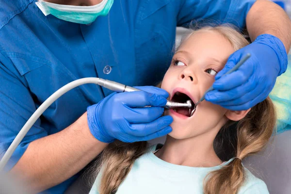 Plan recadré de dentiste avec pièce à main dentaire travaillant avec des dents de petit enfant — Photo de stock