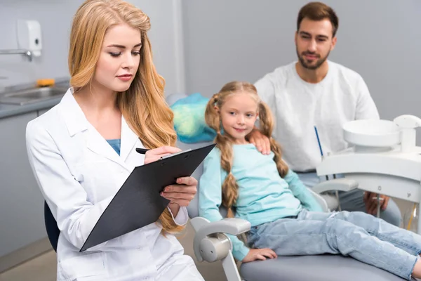 Atractiva mujer dentista escritura en portapapeles mientras que el niño pequeño y su padre sentado en el fondo - foto de stock