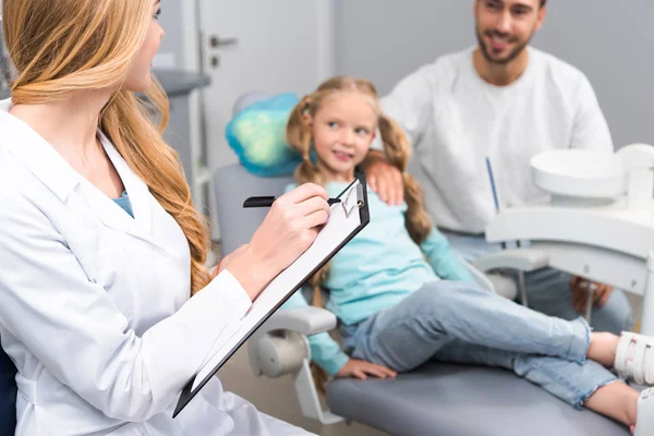 Female dentist with clipboard talking to little child and her young father — Stock Photo