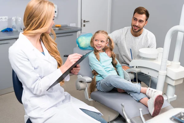 Joven dentista escribiendo en portapapeles, mientras que el niño y su padre sentado en el fondo - foto de stock