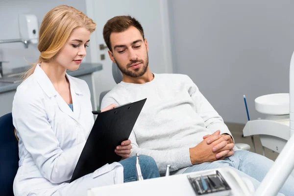 Beautiful female dentist with clipboard and client sitting in dentist office — Stock Photo