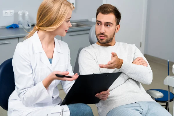 Joven dentista con portapapeles y cliente hablando entre sí en el consultorio del dentista - foto de stock