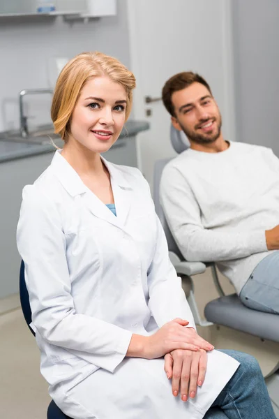 Щаслива жінка-стоматолог і красивий молодий клієнт дивиться на камеру в стоматологічному кабінеті — стокове фото