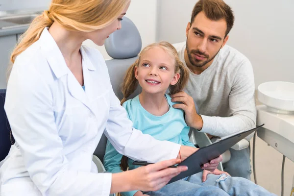 Young female dentist showing clipboard with diagnosis to father and little daughter — Stock Photo