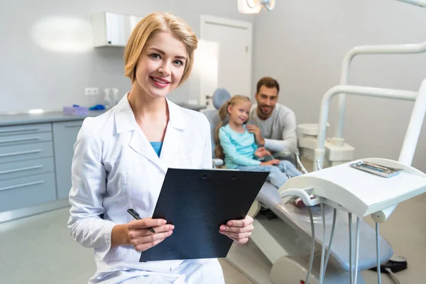 Bela dentista feminina com área de transferência olhando para a câmera enquanto criança pequena e seu pai sentado no fundo — Fotografia de Stock