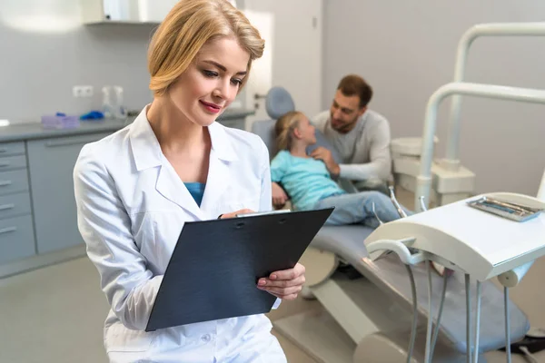 Female dentist writing in clipboard while little child and her father sitting on background — Stock Photo