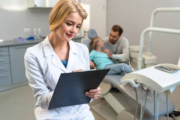 Dentista mujer escribiendo en portapapeles, mientras que el niño pequeño y su padre sentado en el fondo - foto de stock