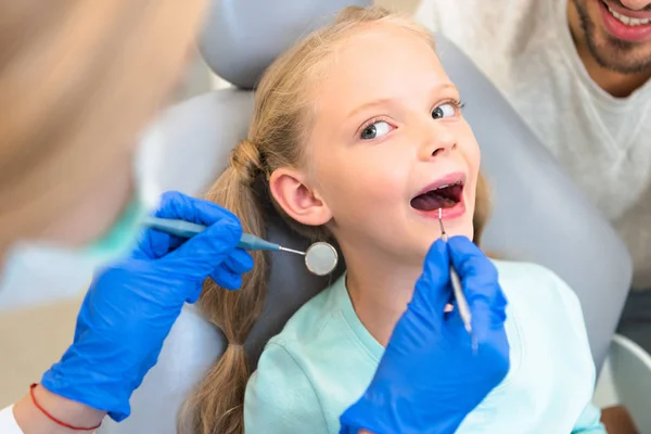 Recortado tiro de mujer dentista examinando dientes de niño pequeño mientras padre sentado cerca de ella - foto de stock