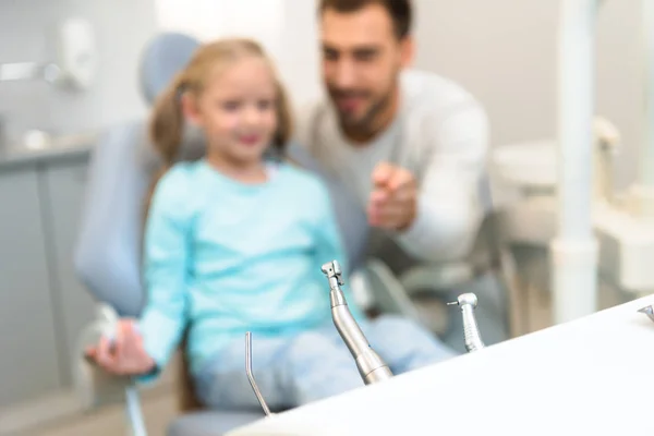 Primer plano de herramientas de dentista en el stand con el padre borroso y la hija en el fondo - foto de stock