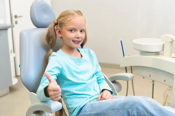 Beau petit enfant assis dans la chaise dentaire au cabinet du dentiste et montrant pouce vers le haut — Photo de stock