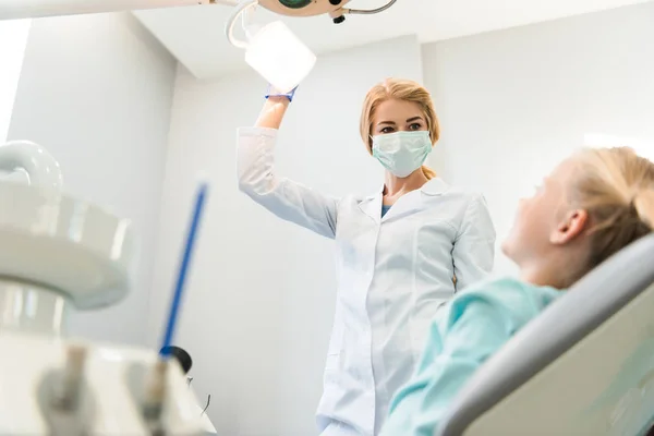 Vue du bas de la jeune dentiste avec lampe debout sur le petit enfant dans la chaise — Photo de stock