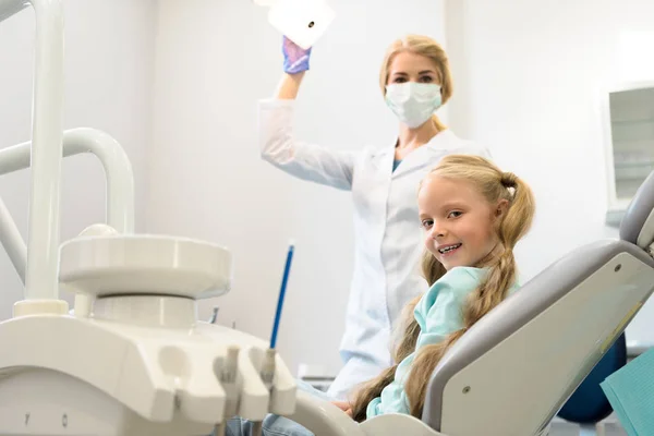 Glückliches kleines Kind blickt in die Kamera, während es im Stuhl in der Zahnarztpraxis sitzt — Stockfoto