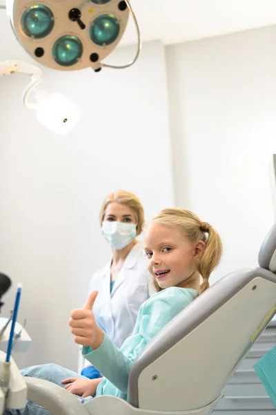Lächelndes kleines Kind zeigt Daumen nach oben, während es im Stuhl in der Zahnarztpraxis sitzt — Stockfoto