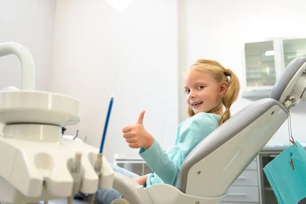 Joyeux petit enfant assis dans une chaise dentaire au cabinet du dentiste et montrant pouce en l'air — Photo de stock