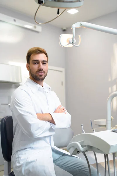 Beau jeune dentiste avec les bras croisés regardant la caméra dans le bureau — Photo de stock