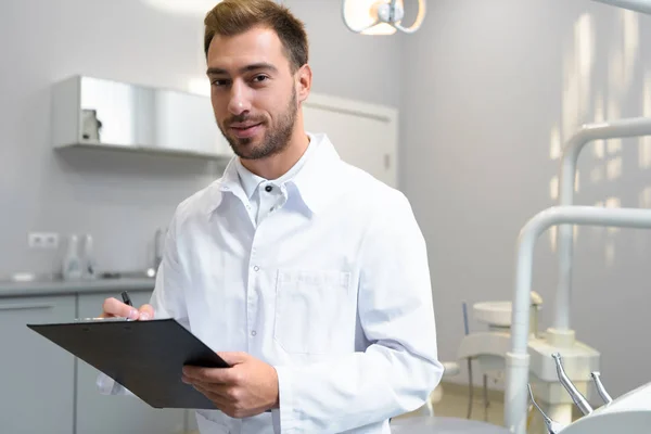 Handsome young dentist in white coat writing in clipboard and looking at camera in office — Stock Photo