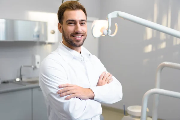 Handsome young dentist with crossed arms looking at camera in office — Stock Photo