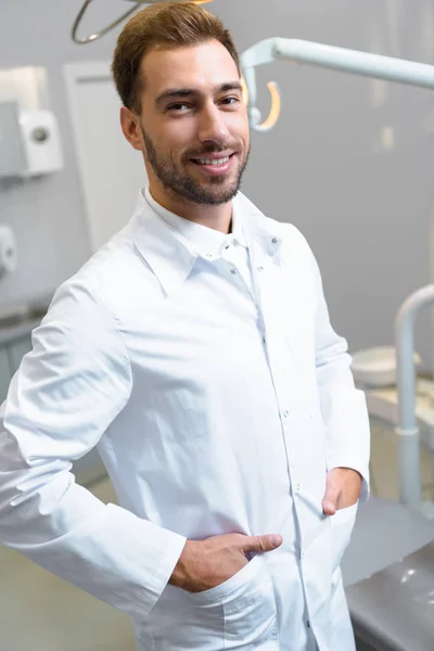 Schöner junger Zahnarzt im weißen Mantel blickt im Büro in die Kamera — Stockfoto