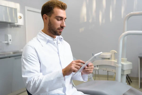 Guapo joven dentista en abrigo usando tableta en la oficina - foto de stock