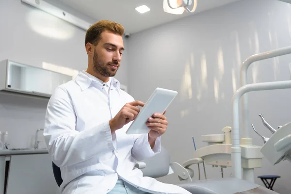 Вид знизу привабливого молодого стоматолога в пальто, використовуючи планшет в офісі — стокове фото