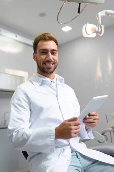 Happy young dentist in coat using tablet in office — Stock Photo