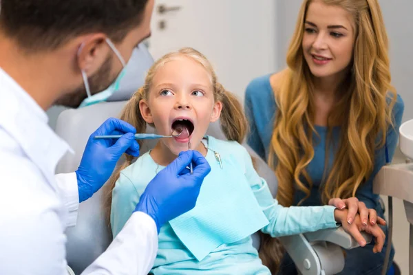 Dentista examinando dentes de criança pequena, enquanto a mãe sentada perto dela — Fotografia de Stock
