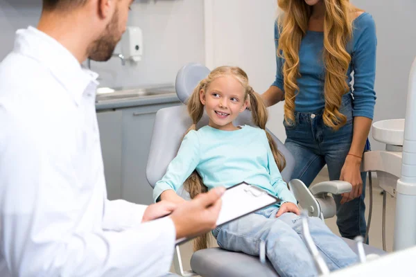 Dentist with clipboard talking with little child while mother standing near her for support — Stock Photo