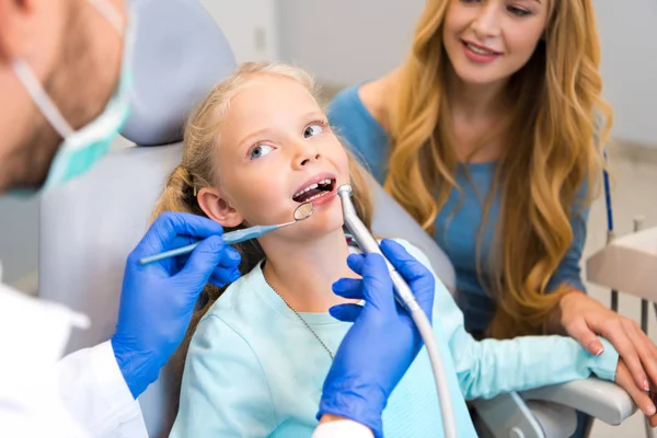 Plan recadré de dentiste travaillant avec un petit enfant tandis que la mère assis près d'elle pour le soutien — Photo de stock