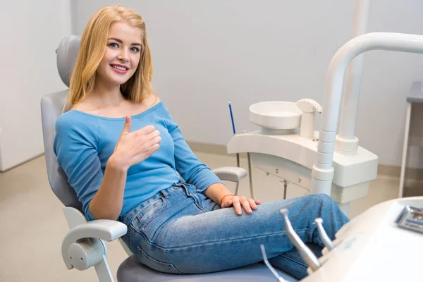 Heureuse jeune femme assise dans la chaise dentaire au cabinet du dentiste et montrant pouce vers le haut — Photo de stock