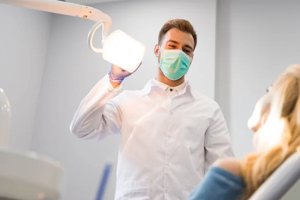 Dentiste en manteau et masque debout sur le client avec lampe — Photo de stock