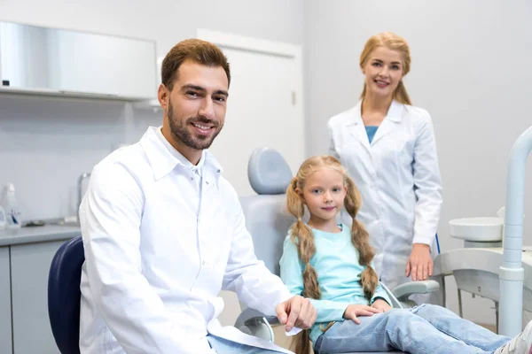 Молодые мужчины и женщины-дантисты с маленьким ребенком, смотрящие в камеру в стоматологическом кабинете — стоковое фото