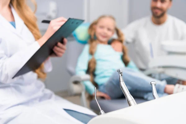 Cropped shot of female dentist writing in clipboard while little child and her young father sitting on background — Stock Photo