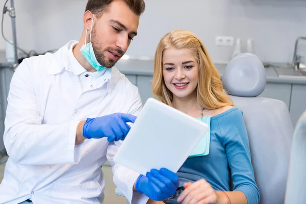 Щасливий молодий стоматолог показує планшет жінці-клієнту в стоматологічному кріслі — стокове фото