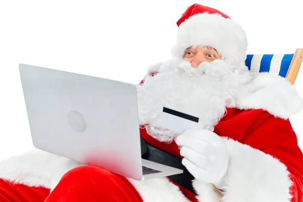 Happy santa claus shopping online with laptop and credit card while sitting on beach chair isolated on white — Stock Photo