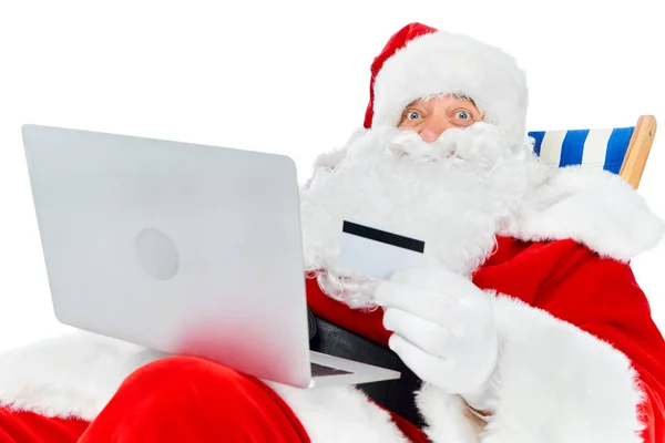 Sorridente Babbo Natale shopping online con computer portatile e carta di credito mentre rilassarsi sulla sedia a sdraio isolato su bianco — Foto stock