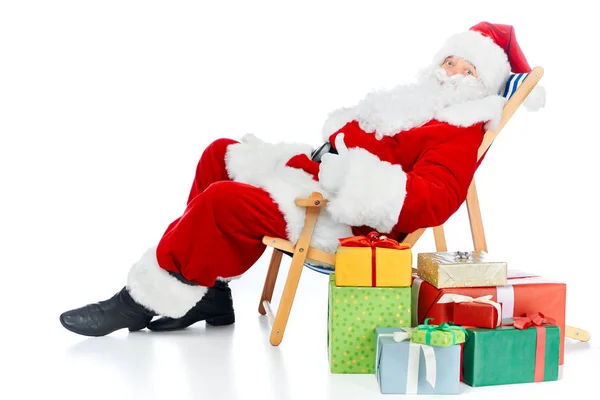 Santa claus mostrando el pulgar hacia arriba mientras descansa en la silla de playa con regalos de Navidad en blanco - foto de stock