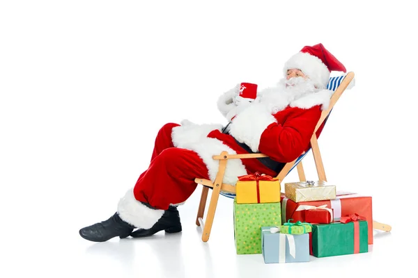 Santa claus holding hot drink and sitting on beach chair with christmas gifts on white — Stock Photo