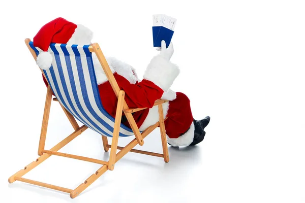 Rear view of santa claus holding two passports and air tickets for christmas trip while sitting in beach chair isolated on white — Stock Photo