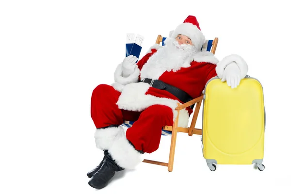 Santa Claus sosteniendo dos pasaportes y billetes de avión para el viaje de Navidad mientras está sentado en la silla de playa con bolsa de viaje aislado en blanco - foto de stock