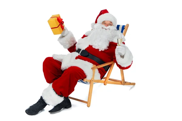 Santa claus holding present and champagne glass while sitting on beach chair isolated on white — Stock Photo