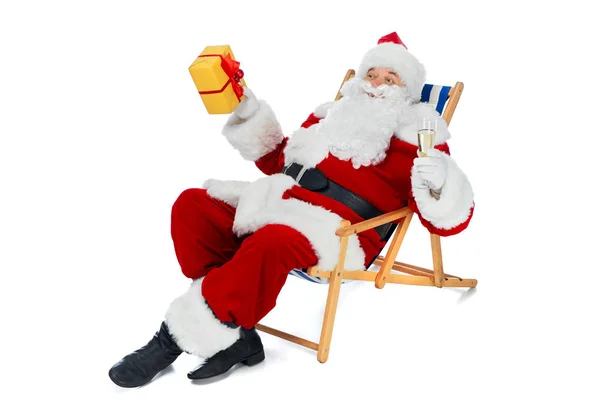Babbo Natale appoggiato sulla sedia da spiaggia con bicchiere di champagne e confezione regalo isolata su bianco — Foto stock