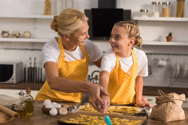 Happy grandmother and granddaughter in aprons smiling each other while preparing cookies together — Stock Photo